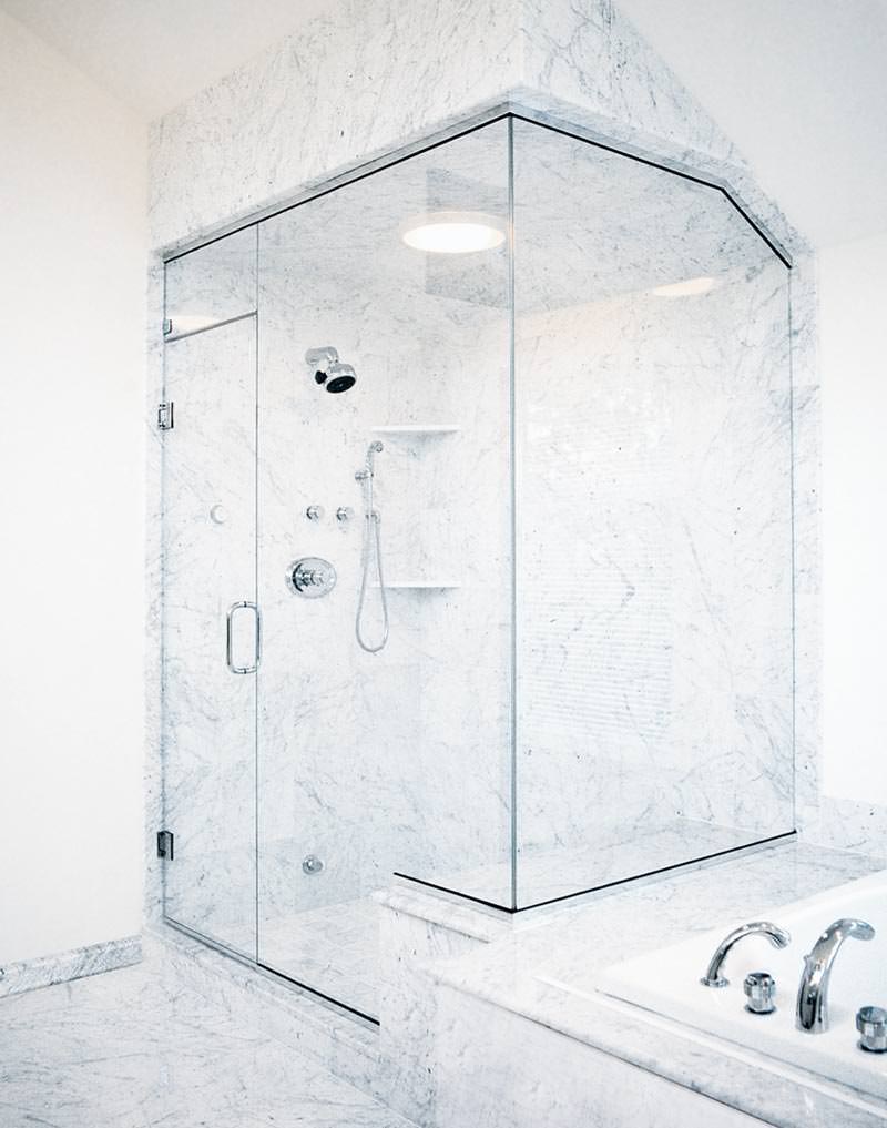 Fully-sealed glass steam enclosure in a corner shower stall surrounded by marble tile with a tubular pull handle.
