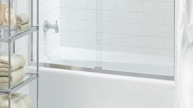 Tub with frameless glass enclosure. Faucet inside the tub and silver glass shelving next to it.